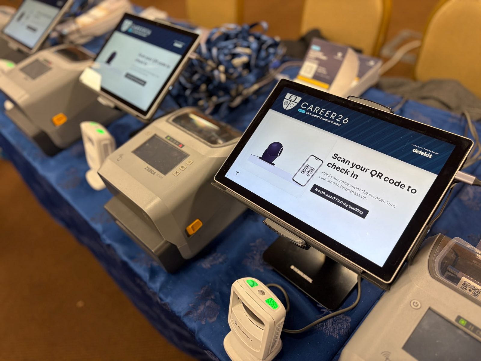 Row of self check-in kiosks at an event, each with a tablet showing a branded 'Scan your QR code to check in' screen, a QR scanner and a badge printer